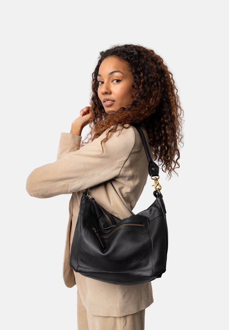 Schwarze Leder-Schultertasche mit einer geschwungenen Form, ausgestattet mit Reißverschlüssen und goldfarbenen Beschlägen, getragen von einer Person in einem beigen Blazer.