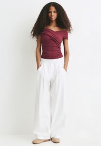 Femme aux cheveux bouclés portant un top bordeaux ajusté à épaules dénudées, un large pantalon blanc avec poches, et des chaussures beiges, se tenant contre un fond blanc.