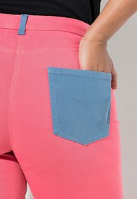 Dopasowane różowe spodnie z kontrastową niebieską kieszenią naszytą. Tkanina wydaje się gładka, z lekkim dodatkiem rozciągliwości dla komfortu.