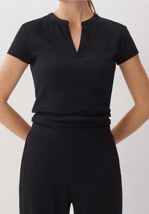 Kobieta ubrana w czarną bluzkę z krótkim rękawem i wycięciem w kształcie litery V oraz czarne spodnie, stojąca z rękami za plecami.