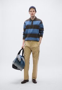 Sinise ja pruuni triibulise kraega polosärk, koorevärvilised püksid ja pruunid kingad. Hoides musta kotti, mille sees on halli ja sinist tekki.