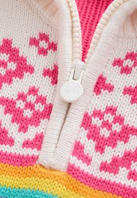 Neulepusero vetoketjulla. Väri pohjana valkoinen, jossa vaaleanpunaisia ja valkoisia kuvioita, sekä monivärinen raidallinen helma oranssin, keltaisen ja petroolin sävyissä.