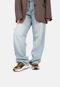 Jeans de pierna ancha de color azul claro combinados con un blazer oversized marrón. Presenta zapatillas gruesas con una mezcla de acentos verdes y marrones.