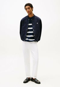 Giacca bomber navy con chiusura a bottoni, camicia a righe navy e bianche sotto, pantaloni bianchi e mocassini neri. Design semplice e moderno.