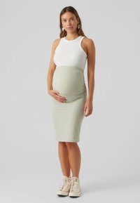 Vestido de maternidade com uma saia justa em verde claro com relevo e uma blusa branca sem mangas, combinado com sapatilhas altas em tom bege.