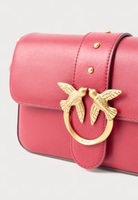 Roza usnjena torbica s zaobljenim poklopcem, zlatni motiv ptice kot zapiralom, dekorativne žebljičke na poklopcu in gladko teksturo.