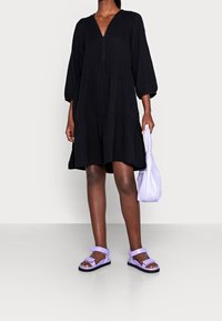 Svart, lager-på-lager klänning med långa ärmar och V-ringning. Bärs med lila sandaler och håller en lavendelfärgad väska. Slät tygstruktur.