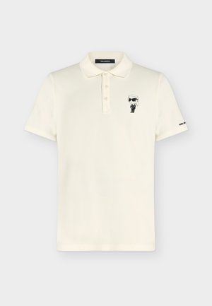 Polo à manches courtes crème avec trois boutons, petite figurine de dessin animé en costume noir et lunettes de soleil brodée sur la poitrine.