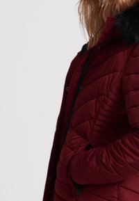 Bordeaux pufferjakke med quiltet design, featuring en faux fur hætte, lynlåslukning og sidelommer. Tekstureret stof med en glat finish.