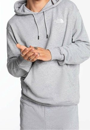 Person trägt einen hellgrauen The North Face Hoodie und passende Hose, die Hände gefaltet und mit mehreren Ringen geschmückt.