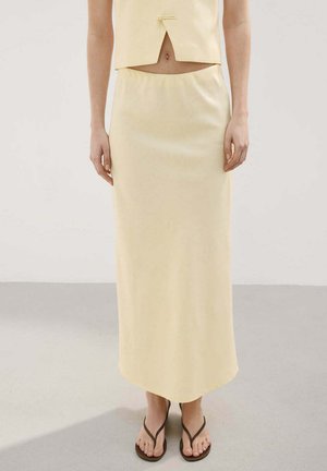 Femme portant une jupe taille haute jaune pâle avec un top court assorti et des sandales marron, debout sur un sol gris.