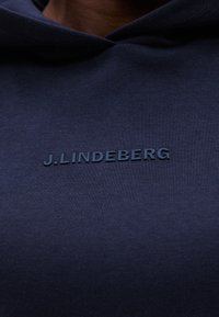 Close-up van marineblauwe stof met verhoogde donkerblauwe tekst "J.LINDEBERG" gecentreerd op het borstgebied.