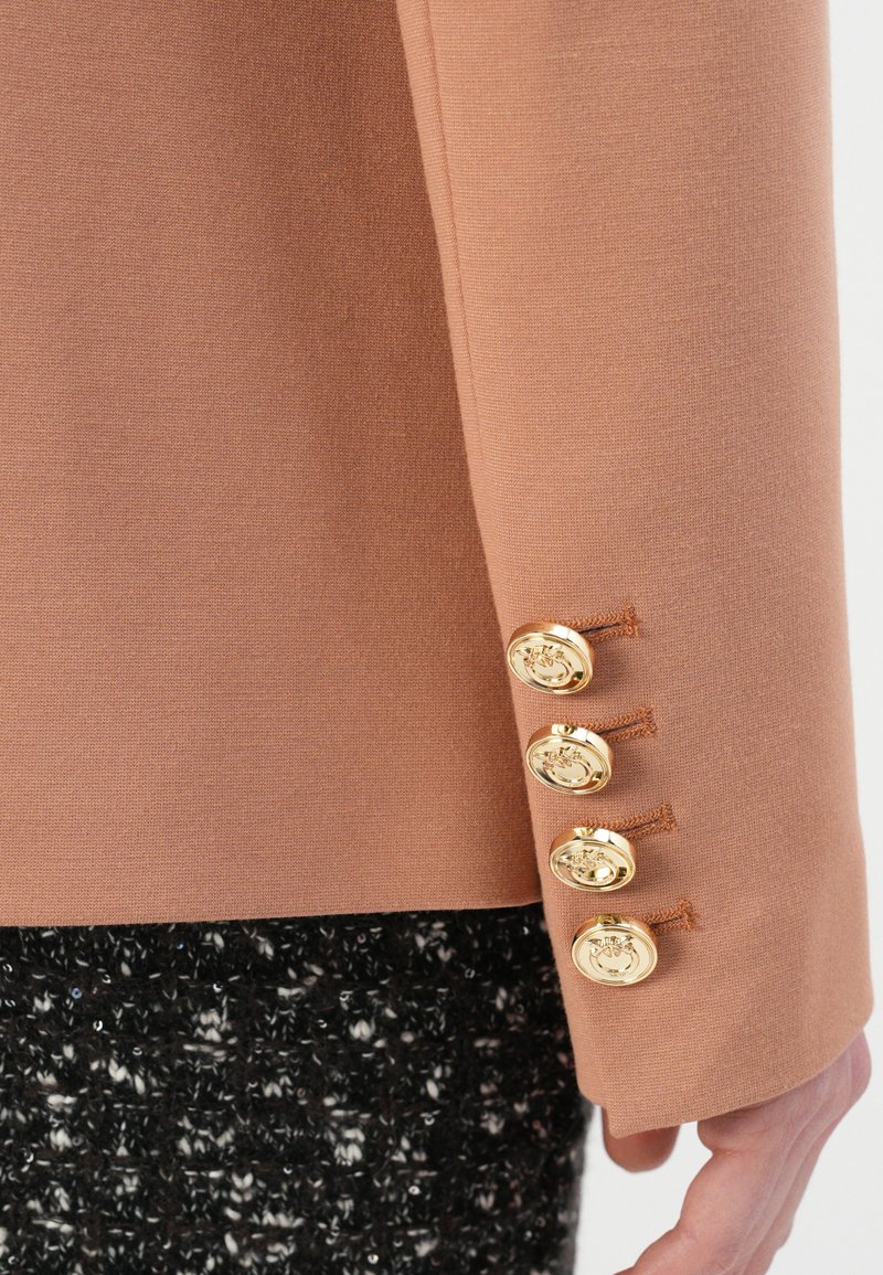 Rękaw w kolorze brzoskwiniowym z czterema złotymi guzikami, o fakturowanej tkaninie i dopasowanym kroju, sięgający czarnej, plecionej odzieży pod spodem.