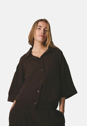 Kobieta o prostych jasnobrązowych włosach nosi luźną ciemnobrązową koszulę na guziki i pasujące spodnie, stoi z rękami w kieszeniach, neutralne tło.