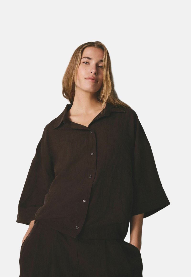 Donna con capelli lisci castano chiaro indossa una camicia marrone scuro morbida con bottoni e pantaloni abbinati, in piedi con le mani nelle tasche, sfondo neutro.