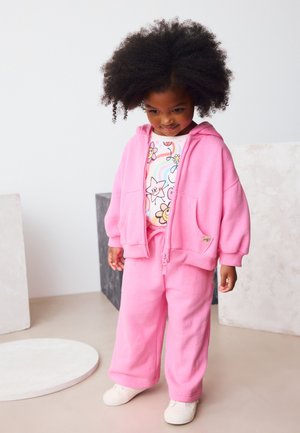 Fiatal gyermek göndör hajjal, rózsaszín kapucnis pulóverben, hozzáillő nadrágban, virágos szivárványos pólóban és fehér cipőben, bent világos háttér előtt áll.