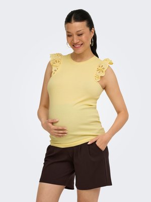 Lachende zwangere vrouw met een geel mouwloos topje met ruches op de schouders en donkere korte broek, één hand op de buik, de andere in de zak.