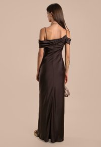 Langes, braunes Satin-Kleid mit schulterfreien Ärmeln, taillierter Corsage und fließendem Rock. Die Accessoires umfassen eine kleine, hellfarbige Handtasche.