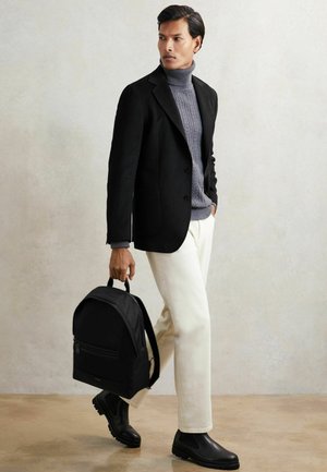 Mann trägt schwarzen Blazer, grauen Rollkragenpullover, weiße Hose, schwarze Stiefel, hält schwarzen Rucksack, geht an einer hellen Wand entlang.