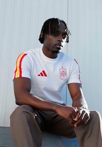 Λευκή ποδοσφαιρική φανέλα με κόκκινες και κίτρινες ρίγες στα μανίκια, με κόκκινο έμβλημα και λογότυπο Adidas. Συνδυάζεται με μπεζ παντελόνια.