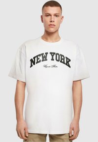 Baltos medvilnės marškinėliai su atsipalaidavusiu siluetu, su juodu „NEW YORK“ spaudiniu ir „Upgrade Studios“ užrašu kursyvu žemiau. Minkšta tekstūra ir trumpomis rankovėmis.