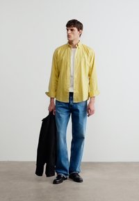 Νέος άνδρας που φοράει κίτρινο πουκάμισο πάνω από λευκό t-shirt, μπλε τζιν, μαύρα παπούτσια, κρατώντας μαύρο μπουφάν, στέκεται μπροστά σε απλή λευκή τοίχο.