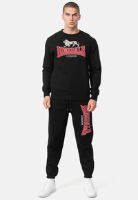 Felpa nera con logo rosso e bianco, collo a girocoscia, polsini a costine, abbinata a pantaloni jogger neri con un design di logo simile.