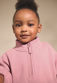 Rožinės spalvos megztinis su užtrauktuku, aukštu apykakliu ir trumpomis rankovėmis, pagamintas iš minkštos medžiagos. Paprastas dizainas be matomų raštų ar akcentų.