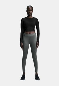 Schwarzes langärmliges Crop-Top mit rundem Ausschnitt, kombiniert mit eng anliegenden, hoch taillierten, olivgrünen Leggings mit Reißverschlusstaschen. Schwarze Sneakers.
