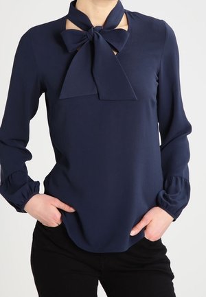 Donkerblauwe blouse met lange mouwen en een grote gestrikte strik bij de hals, gedragen in een zwarte broek, handen in de zakken.