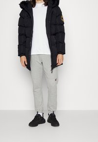 Chaqueta de plumas negra con capucha y ribete de pelo, camiseta blanca de manga larga, pantalones de chándal grises y zapatillas negras. Las características incluyen cremalleras y parches.