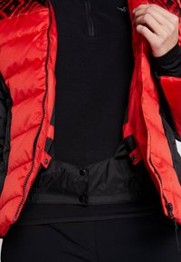 Personne portant une doudoune rouge dézippée sur un haut en polaire noire, tenant la veste ouverte pour montrer les détails intérieurs.