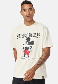 Camiseta de algodón beige con un gráfico de estilo vintage de Mickey Mouse en pantalones cortos rojos. Manga corta y corte relajado. Etiqueta de logo negra en el dobladillo.