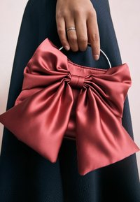 Punainen satiinilaukku, jossa on suuri nauhadesign, metallinen kahva ja teksturoitu pinta. Nauhassa on pehmeät taitteet ja keskimmäinen solmu.