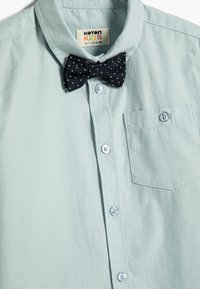 Camisa de manga corta azul claro hecha de algodón, con un bolsillo en el pecho izquierdo, cierre de botones en el frente y una pajarita de lunares negros sujeta al cuello.