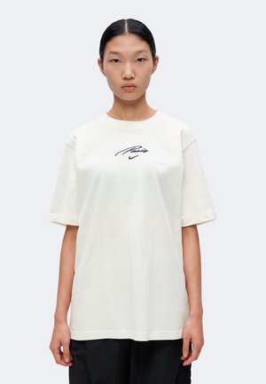 Jeune femme aux cheveux noirs raides porte un t-shirt blanc oversize avec l'inscription « Paris » et un pantalon noir sur un fond uni.