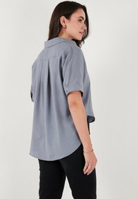 Camicia grigia chiara a maniche corte con piega posteriore, polsini abbottonati e orlo arrotondato. Texture liscia e vestibilità comoda.