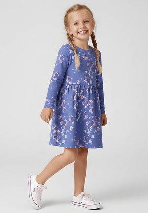 Uśmiechnięta młoda dziewczyna z warkoczami, ubrana w niebieską sukienkę z długim rękawem ozdobioną różowymi kwiatowymi wzorami oraz białe trampki, stojąca na jednej nodze.