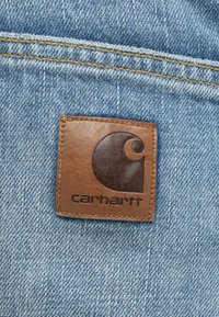 Barna bőrfolt dombornyomott logóval, kék farmer anyagra ragasztva; sárga varrással és texturált háttérrel.