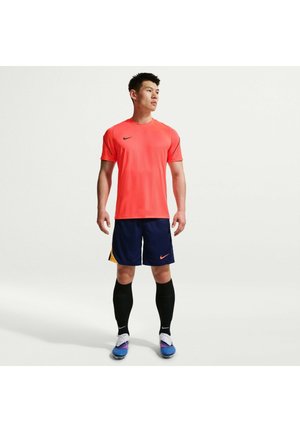 Homme debout portant un t-shirt rouge vif Nike, un short bleu marine avec des accents orange, des chaussettes noires montantes et des chaussures de football bleues.