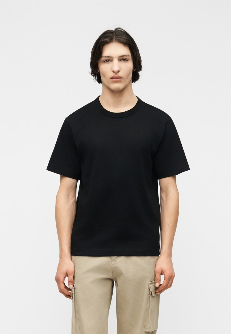 Jeune homme aux cheveux bruns mi-longs portant un t-shirt noir uni à manches courtes et un pantalon cargo beige, debout devant un fond blanc uni.