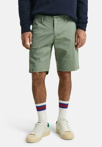 Grønne shorts med rett passform, stofftekstur; kombinert med kremfargede sneakers og hvite sokker med røde og blå striper.