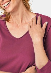 T-Shirt mit V-Ausschnitt aus tiefviolettem Stoff, mit kurzen Ärmeln und einer weichen, glatten Textur. Haut sichtbar am Hals und an der Hand, zeigt natürliche Töne.