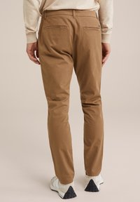 Hnědé bavlněné kalhoty s úzkým střihem, se dvěma zadními kapsami a hladkou texturou, v kombinaci se světlými sportovními botami.