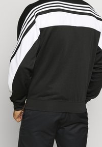 Zwarte sportjack met een wit paneel en drie witte strepen over de schouders. Gemaakt van gladde stof met een ontspannen pasvorm.