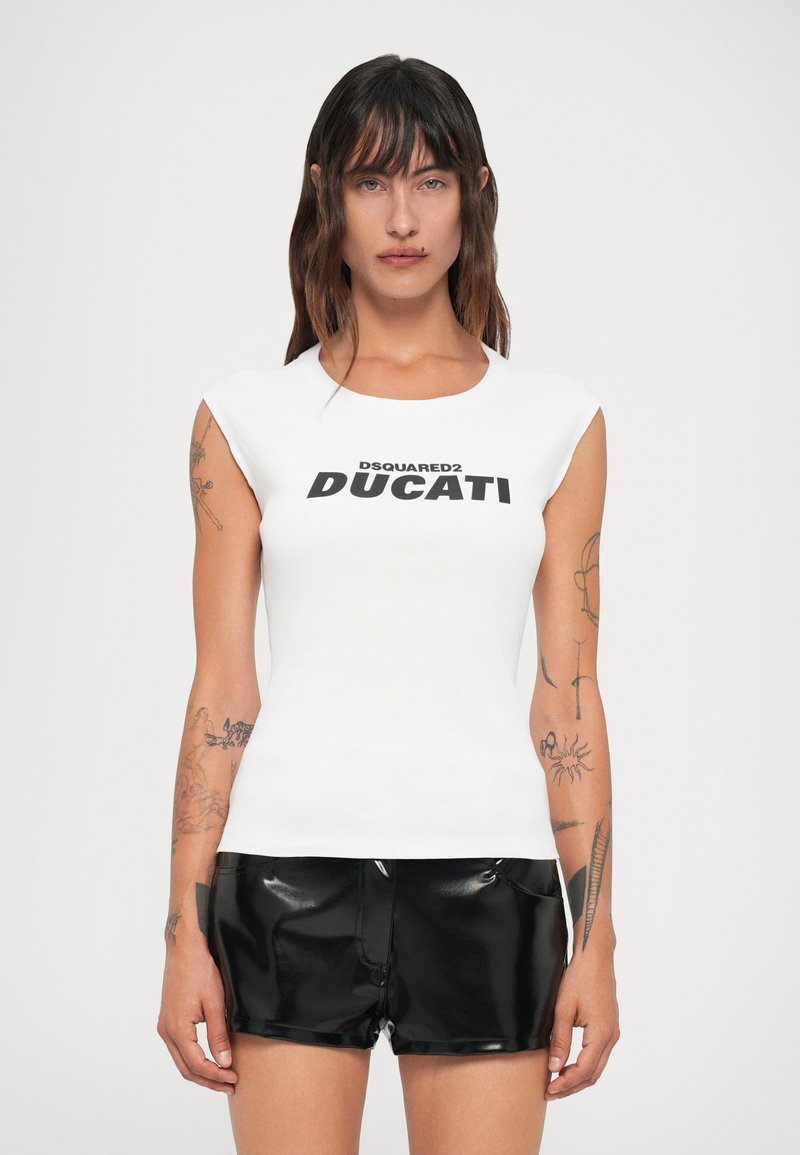 Ženska z dolgimi rjavimi lasmi, oblečena v belo brezrokavno majico DSQUARED2 Ducati in črne usnjene kratke hlače, stoji proti enobarvni ozadju.