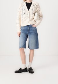 Stickad kavaj i krämfärg med blommönstrad spets och beige knappar, kombinerad med ljus denim bermudashorts, vita strumpor och svarta loafers.