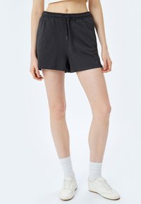 Zwarte, rekbare shorts met een elastische tailleband, trekkoorddetails en een ontspannen pasvorm; gecombineerd met witte sokken en lichte schoenen.
