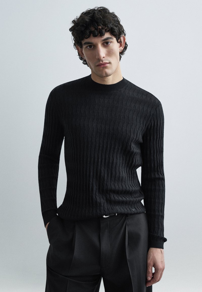 Μαύρο πλεκτό πουλόβερ με ριμπ ύφανση, στρογγυλή λαιμόκοψη και μακριά μανίκια, με υφή στο σχέδιο. Φοριέται με μαύρο παντελόνι κατά παραγγελία.