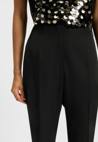 Schwarze Hosen mit hohem Bund, gefaltetem Vorderteil und glatter Textur. Das Oberteil ist mit glänzenden, runden Pailletten in Schwarz und Gold verziert.